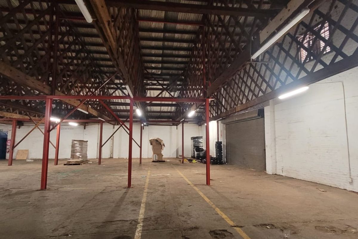 Spacious industrial warehouse featuring high exposed timber truss ceilings, steel support columns, roller shutter door access, and painted brick walls.