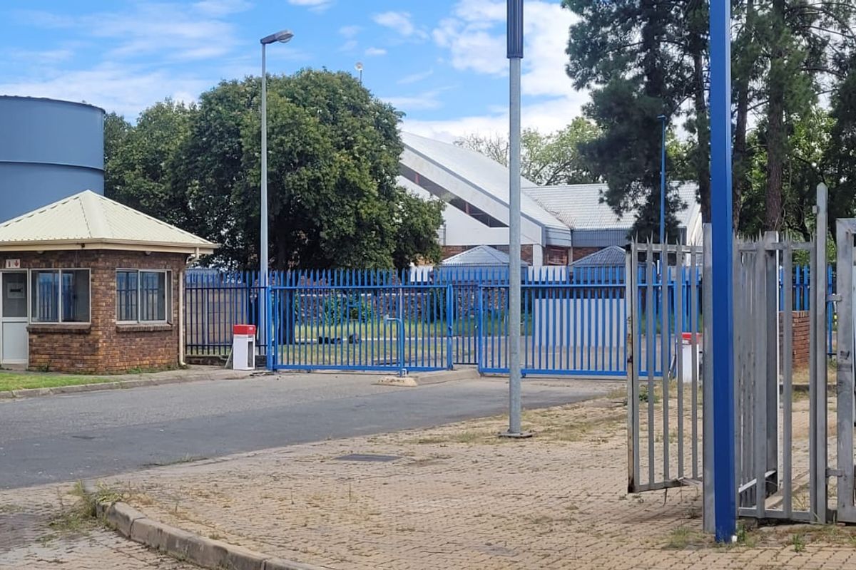 Industrial property entrance featuring a security guard house, blue metal perimeter fencing, paved driveway, and large storage tanks.