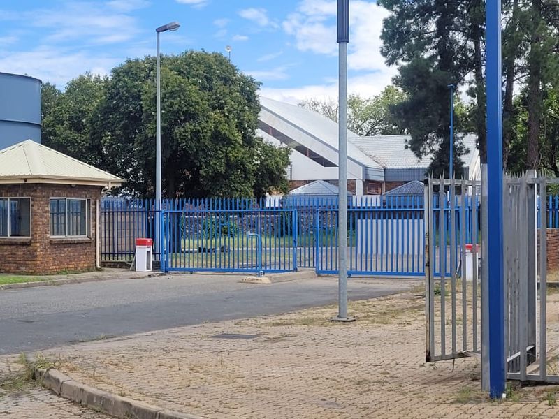 Industrial property entrance featuring a security guard house, blue metal perimeter fencing, paved driveway, and large storage tanks.