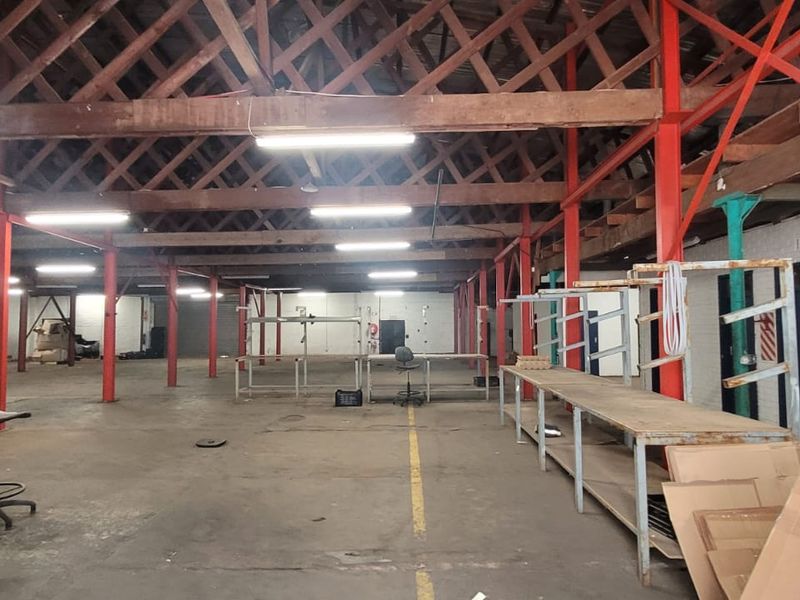 Spacious industrial warehouse featuring exposed timber roof trusses, red painted steel support columns, concrete flooring, and integrated fluorescent lighting throughout the open floor plan.