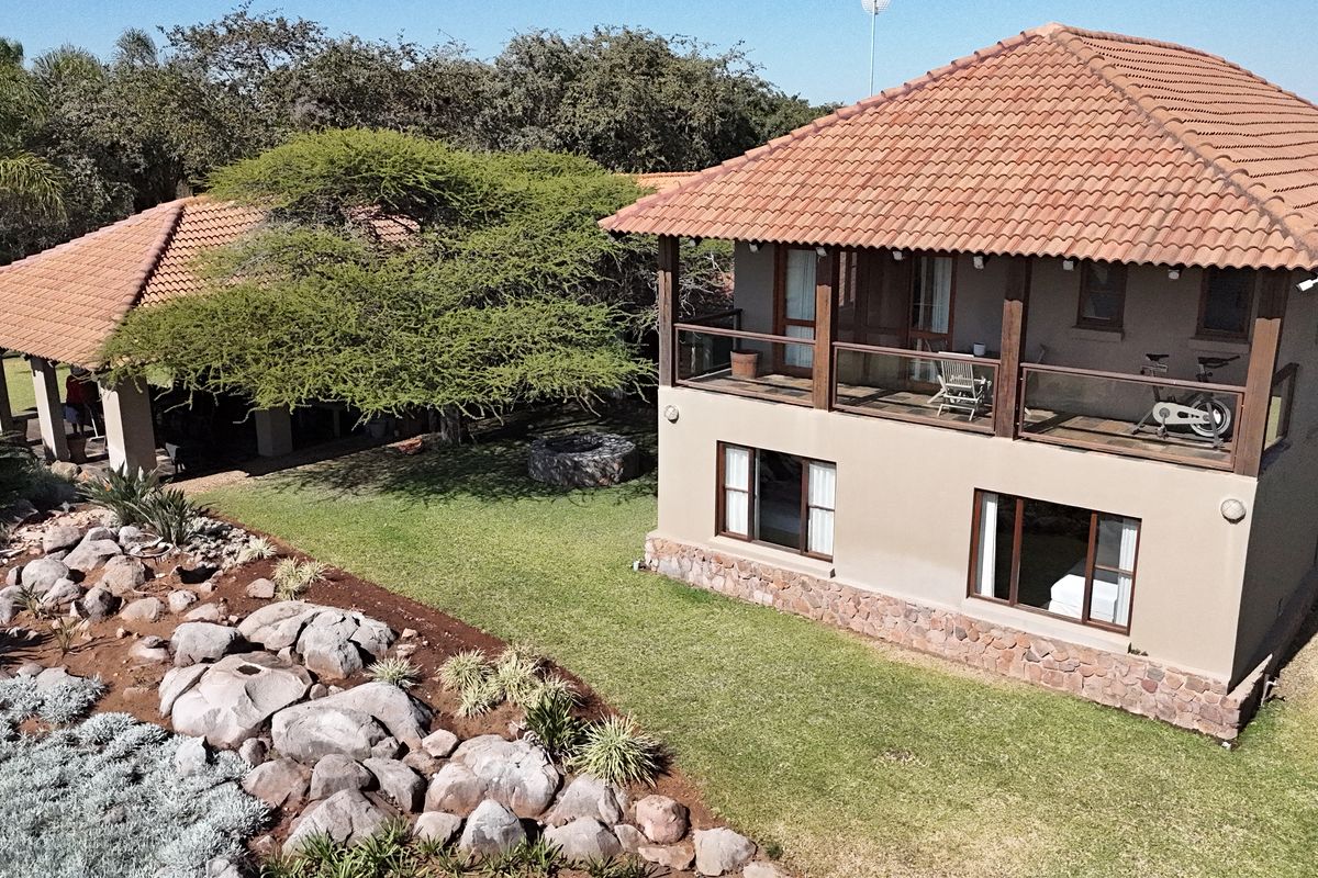 Aerial view of a two-story farm residence featuring terracotta roofing, stone cladding, and a spacious upper-level balcony overlooking a landscaped garden.