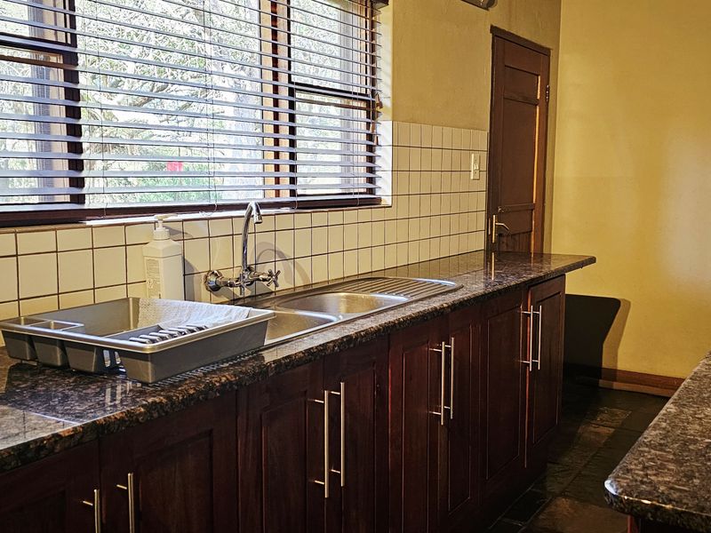 A functional kitchen area featuring dark wooden cabinetry, granite countertops, a double basin sink, and a tiled backsplash with natural light.