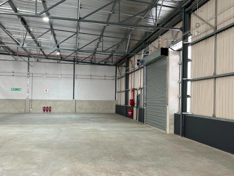 Spacious industrial warehouse unit featuring high ceilings with exposed steel trusses, concrete flooring, and a large roller shutter door for easy access.