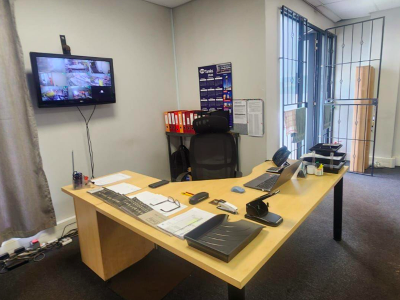 A functional industrial office space featuring a drop ceiling, security gate, and wall-mounted CCTV monitoring system.