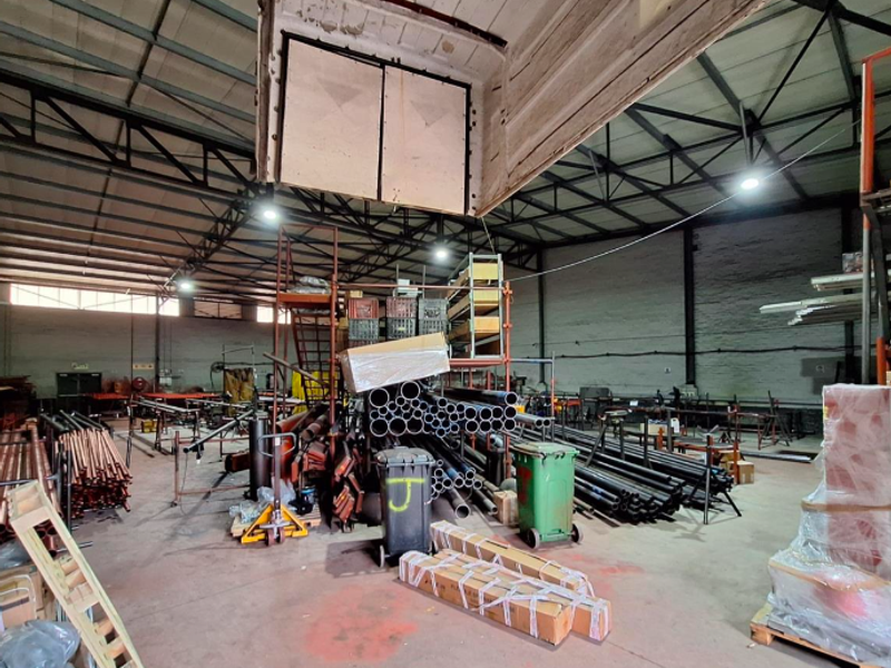 Spacious industrial warehouse area featuring high exposed steel roof trusses, concrete flooring, and brick walls, currently utilized for storage and industrial operations.