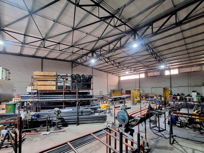 Spacious industrial warehouse facility featuring high-clearance exposed steel roof trusses, concrete flooring, and high-bay lighting, currently utilized for metal fabrication and storage.