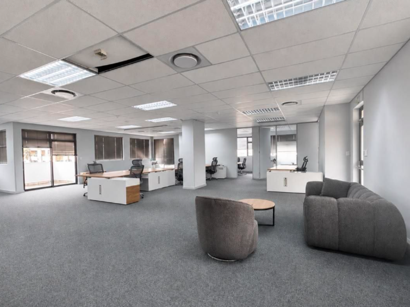 Spacious open-plan office area featuring a suspended ceiling, recessed lighting, carpeted flooring, and large windows with blinds.
