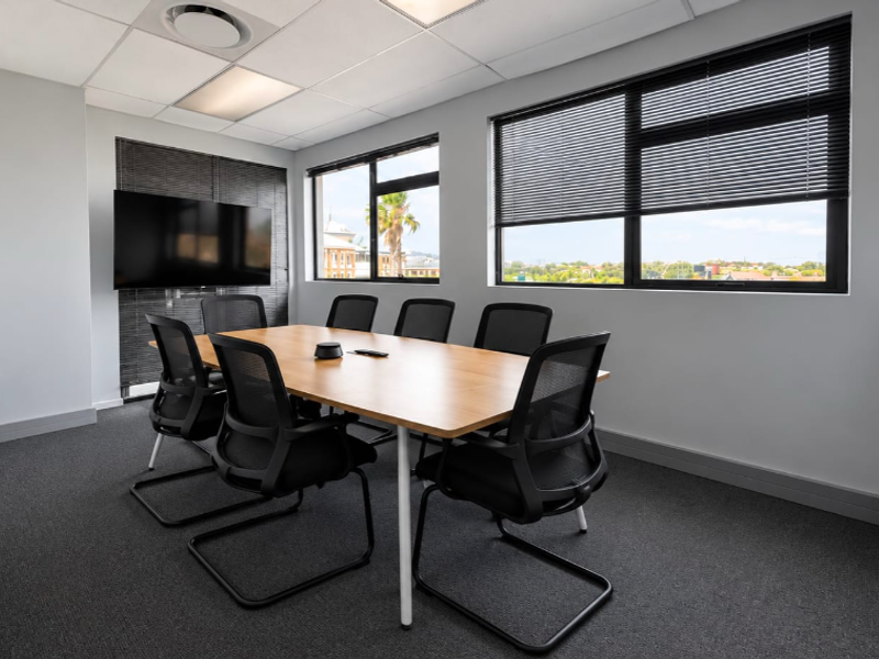 A modern, well-lit office meeting room featuring neutral finishes, carpeted flooring, and large windows with venetian blinds providing natural light and exterior views.