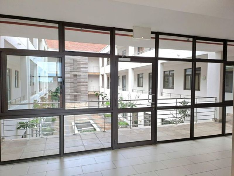A bright, modern office hallway featuring extensive aluminium-framed glass partitioning and tiled flooring, overlooking an internal courtyard.