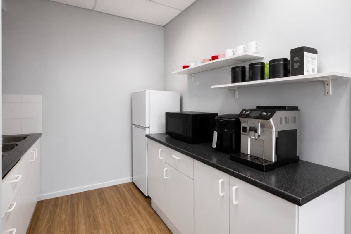 A clean and functional office kitchenette featuring white cabinetry, dark laminate countertops, a stainless steel sink, and wood-look flooring.