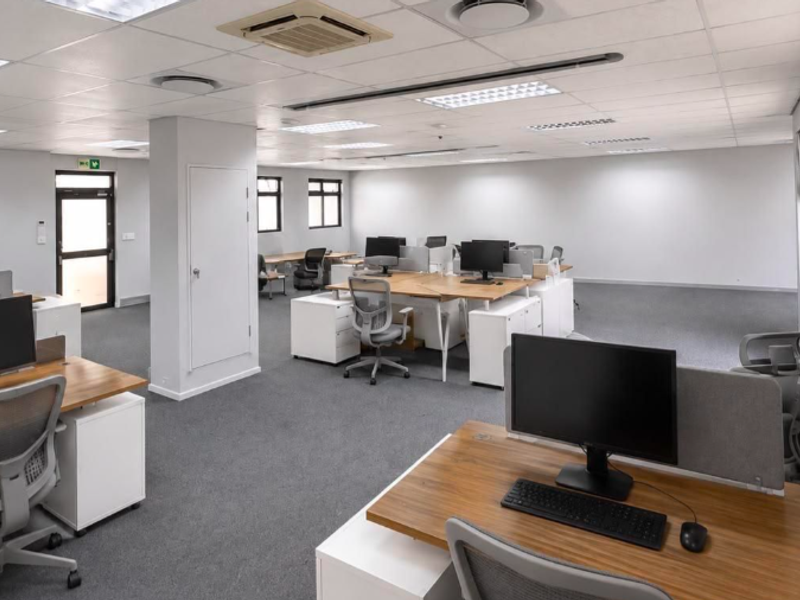 A spacious, well-lit open-plan office area featuring neutral finishes, carpeted flooring, and integrated ceiling-mounted air conditioning.