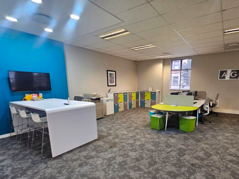 A modern, well-lit office space featuring carpeted flooring, a suspended ceiling with recessed lighting, and integrated storage lockers.