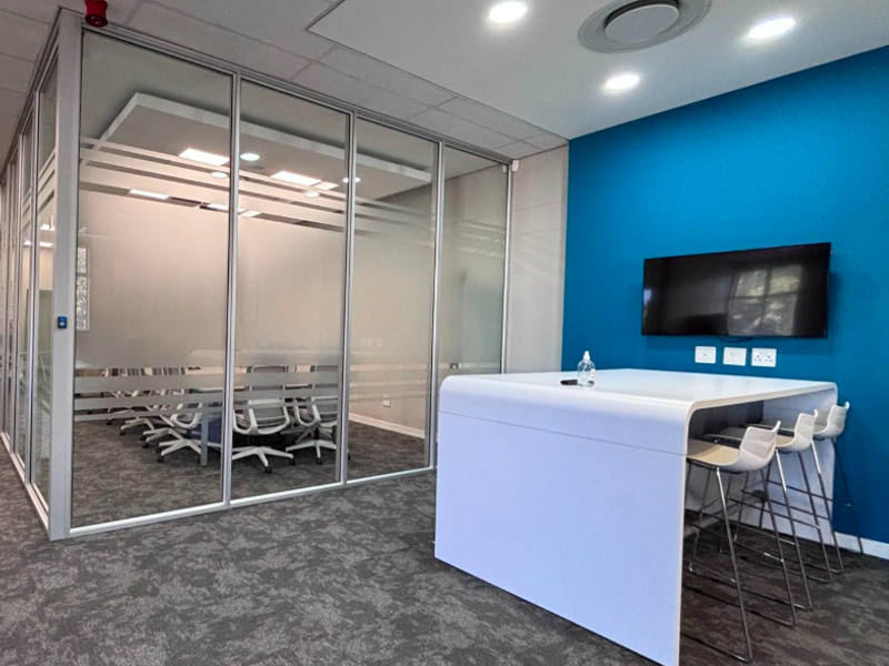 Modern office space featuring a glass-partitioned meeting room, blue accent wall, integrated ceiling lighting, and professional carpeted flooring.