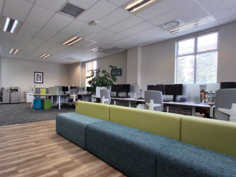 Modern open-plan office space featuring a mix of carpet and wood-look flooring, suspended ceilings with recessed lighting, and large windows providing natural light.
