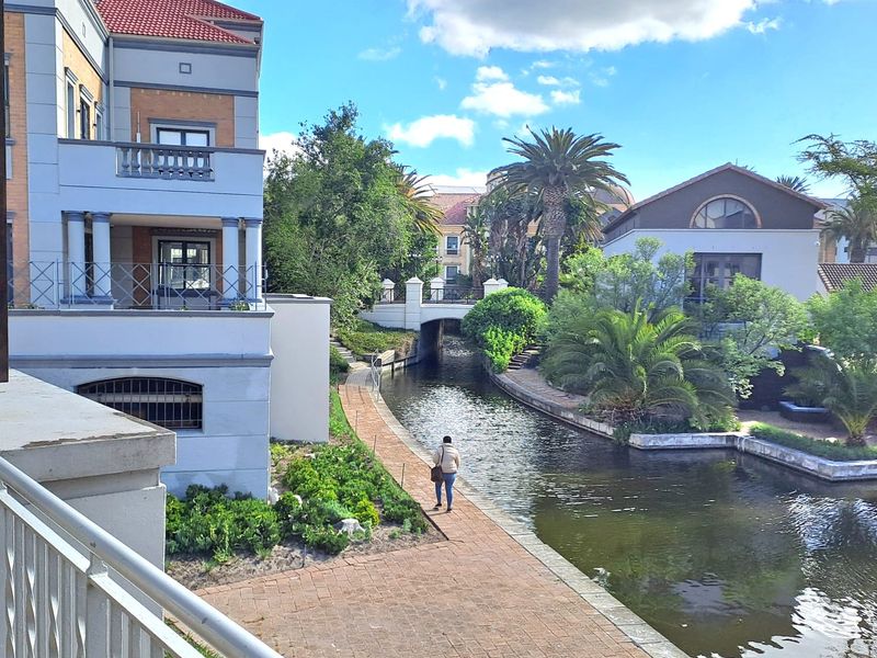 A scenic view of the office property exterior featuring a canal-side paved walkway, landscaped gardens, and a bridge connecting the building complex.