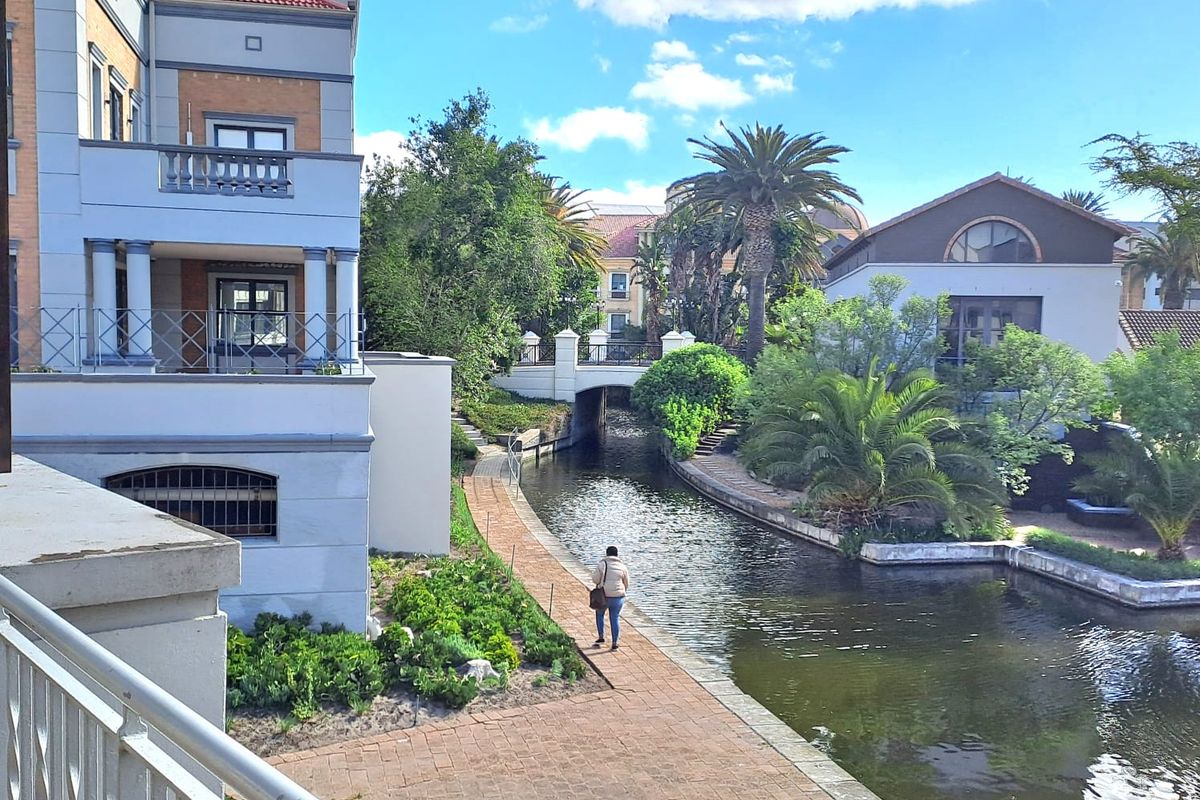 A scenic view of the office property exterior featuring a canal-side paved walkway, landscaped gardens, and a bridge connecting the building complex.