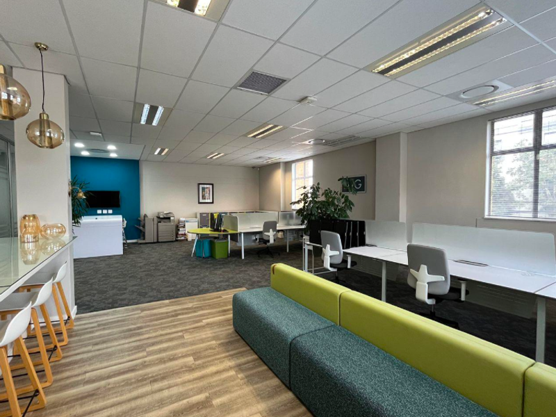 Modern open-plan office space featuring a mix of carpet and wood-look flooring, suspended ceilings, and integrated lighting with a dedicated breakfast bar area.