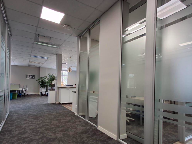 Modern office interior featuring glass partition walls, suspended ceilings with recessed lighting, and professional carpet flooring in a clean, corporate environment.