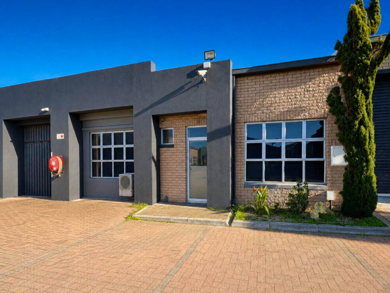 Front elevation of a modern office unit featuring a combination of brick and rendered finishes, paved parking area, and secure roller shutter access.