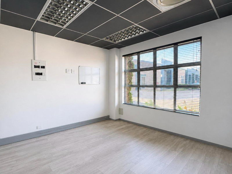 A bright, empty office space featuring modern laminate flooring, a suspended ceiling with recessed lighting, and large windows with fitted venetian blinds.