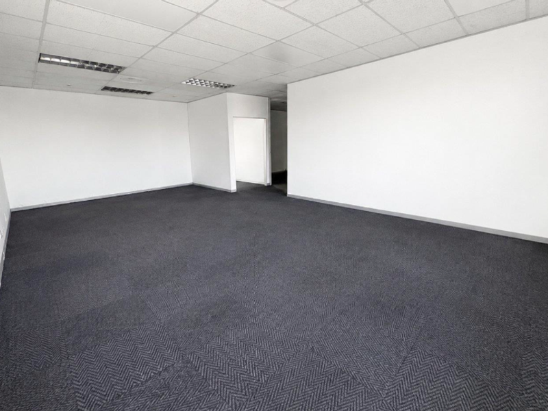A spacious, empty office area featuring a suspended ceiling with recessed lighting and dark grey carpet tiles.