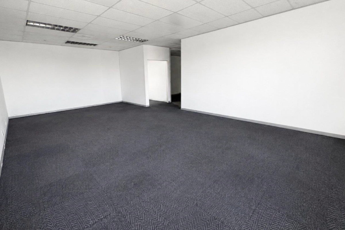 A spacious, empty office area featuring a suspended ceiling with recessed lighting and dark grey carpet tiles.