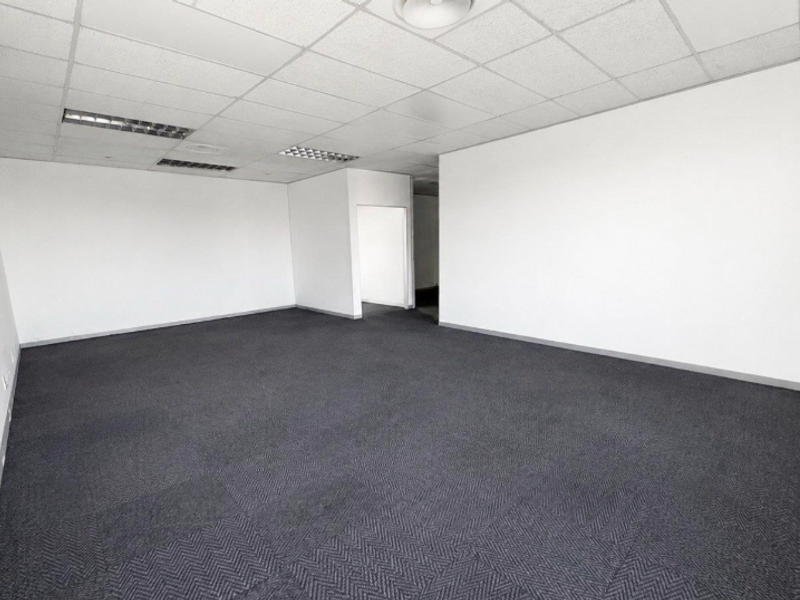 A spacious, empty office area featuring neutral walls, dark carpet tiles, and a suspended ceiling with recessed lighting.