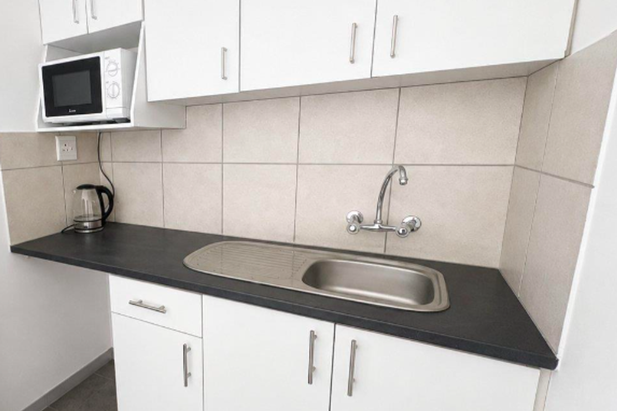 A compact, clean kitchenette featuring white cabinetry, dark laminate countertops, a stainless steel sink, and neutral tiled backsplash.