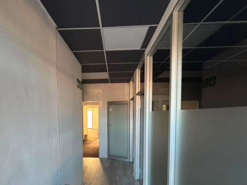 A modern office corridor featuring glass partitions, a dark suspended ceiling, and tiled flooring, providing a clean and professional workspace environment.
