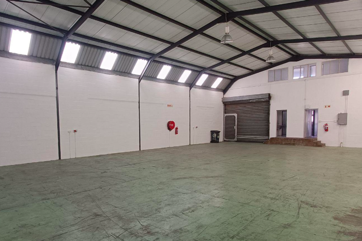 Spacious open-plan office or warehouse area featuring exposed steel trusses, abundant natural light from skylights, and a secure roller shutter door.