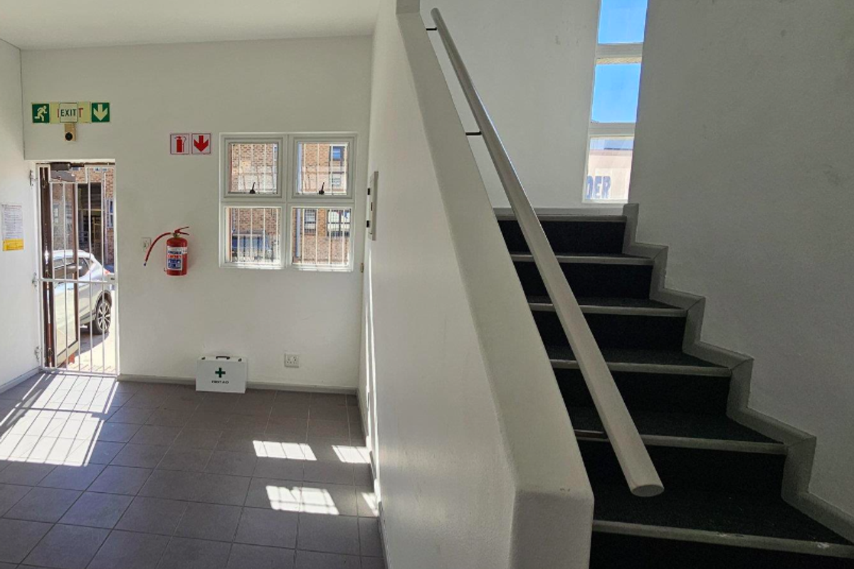 A clean, functional office entrance area featuring a staircase with a handrail, tiled flooring, and essential safety equipment including a fire extinguisher and exit signage.