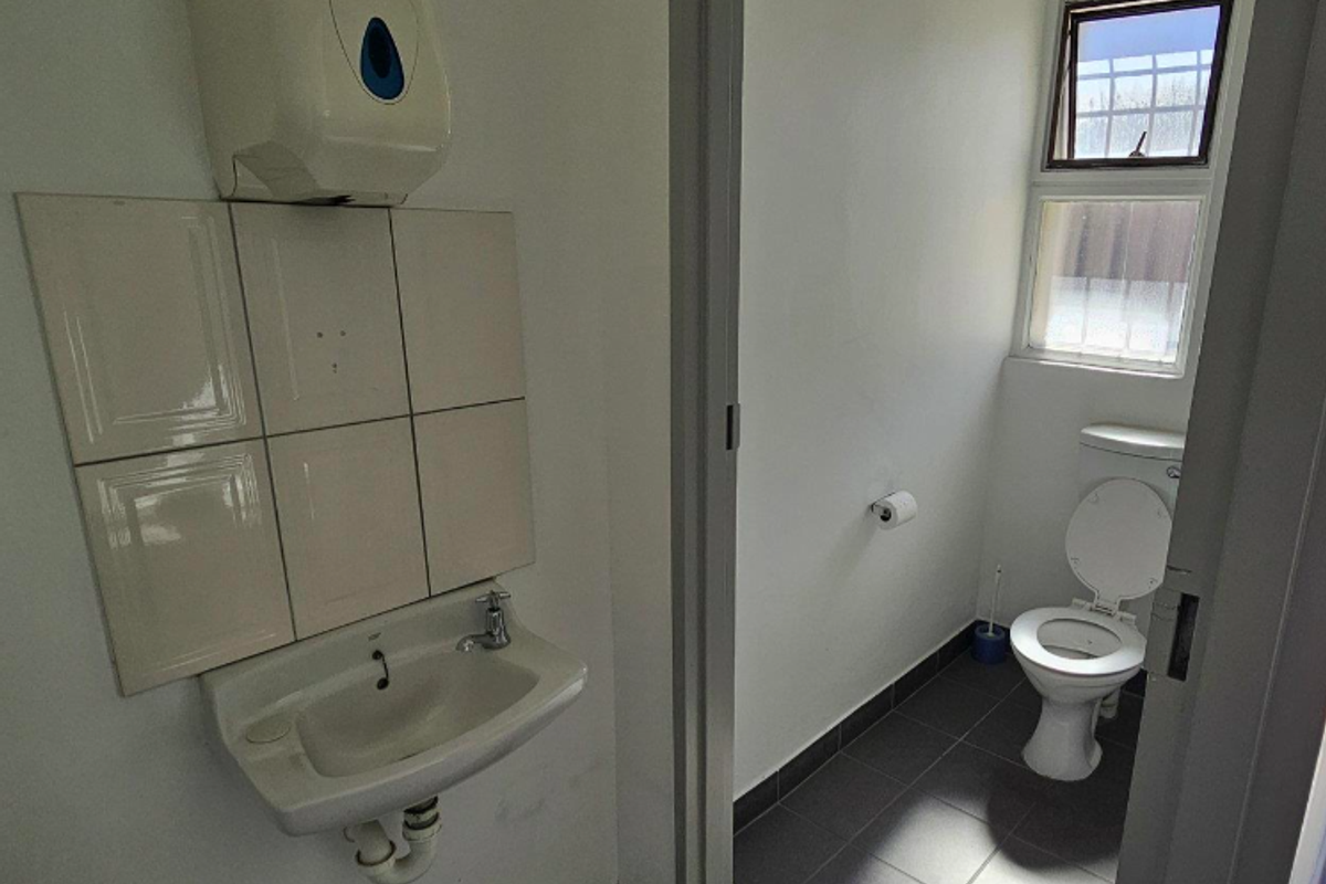 A functional office bathroom featuring a wall-mounted basin with tiled splashback, a standard toilet, and tiled flooring with natural light from a window.
