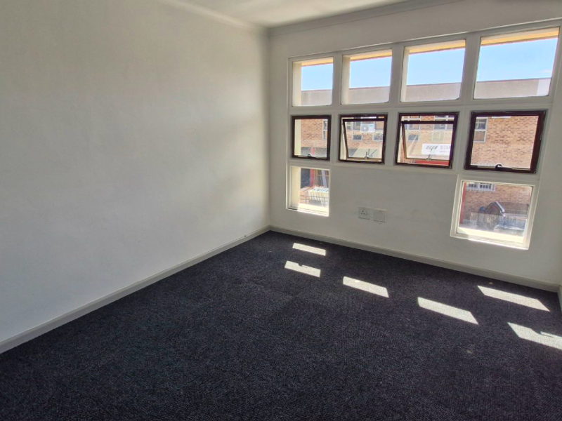 A bright, empty office space featuring dark carpeting, neutral walls, and large multi-pane windows providing natural light.