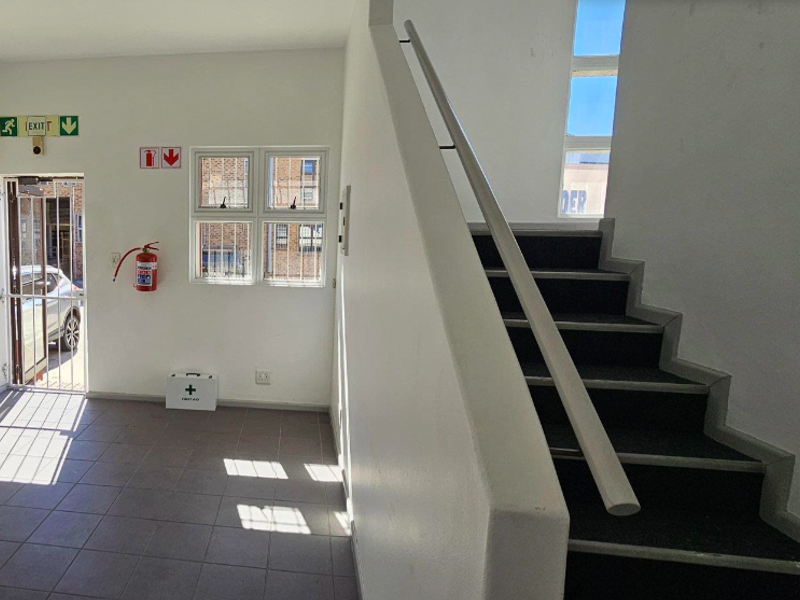 A clean, functional office entrance area featuring a staircase with a handrail, tiled flooring, and essential safety equipment including a fire extinguisher and exit signage.