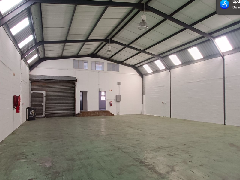 Spacious open-plan office or warehouse area featuring high ceilings with exposed steel trusses, abundant natural light from skylights, and a large roller shutter door.