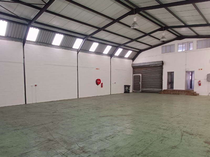 Spacious open-plan office or warehouse area featuring exposed steel trusses, abundant natural light from skylights, and a secure roller shutter door.