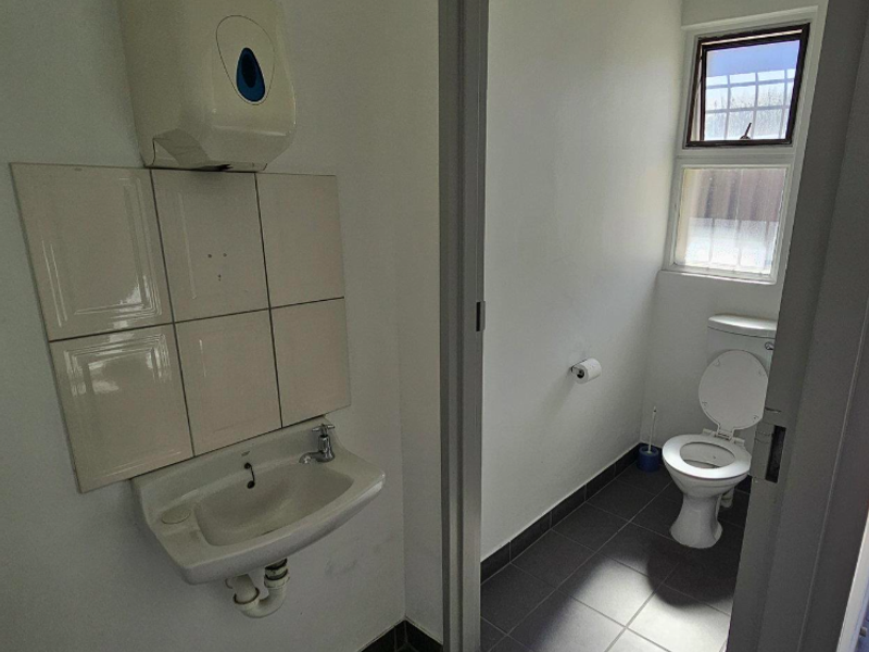 A functional office bathroom featuring a wall-mounted basin with tiled splashback, a standard toilet, and tiled flooring with natural light from a window.