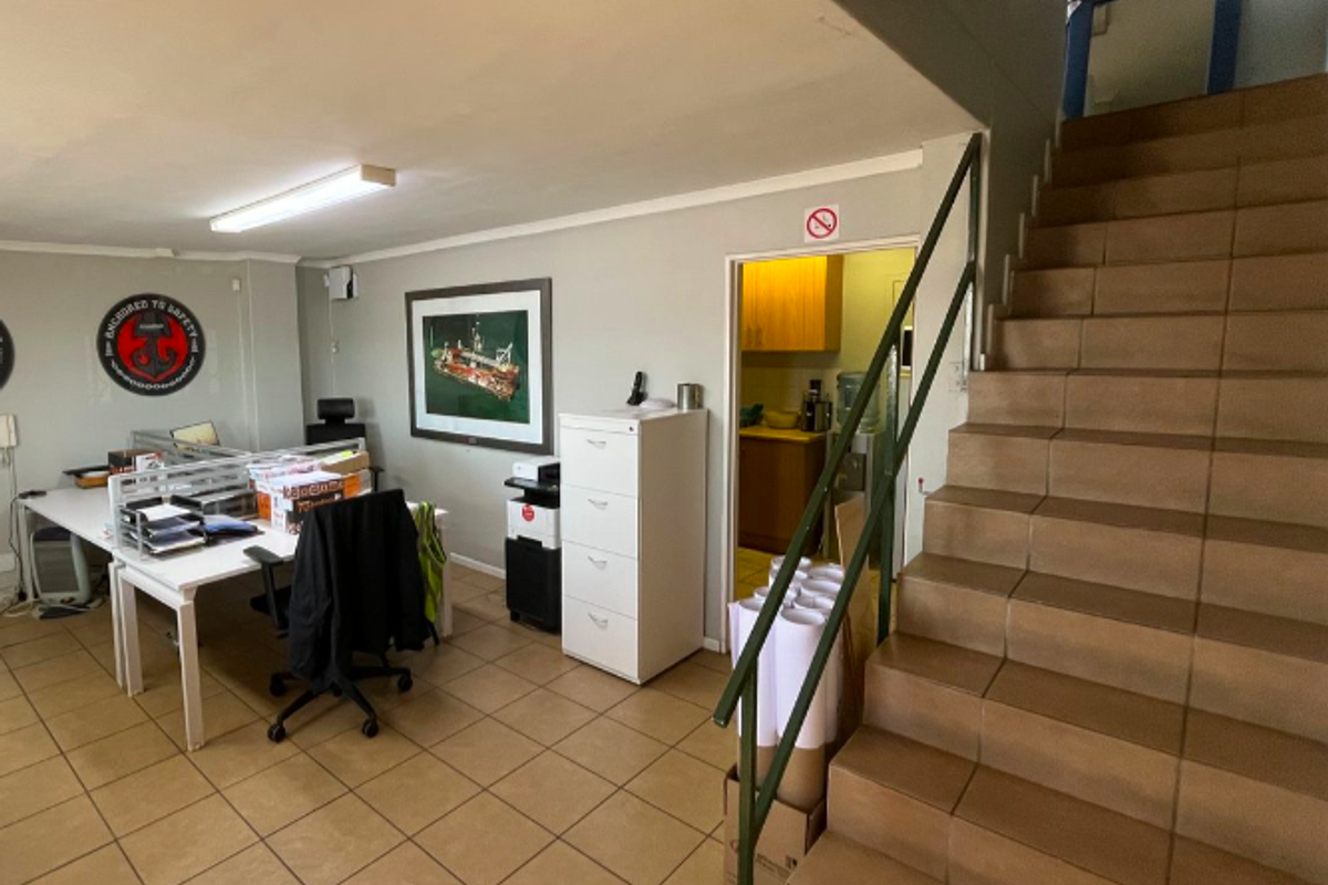 Functional industrial office space featuring tiled flooring, a staircase with a metal handrail, and direct access to an adjacent utility or kitchen area.