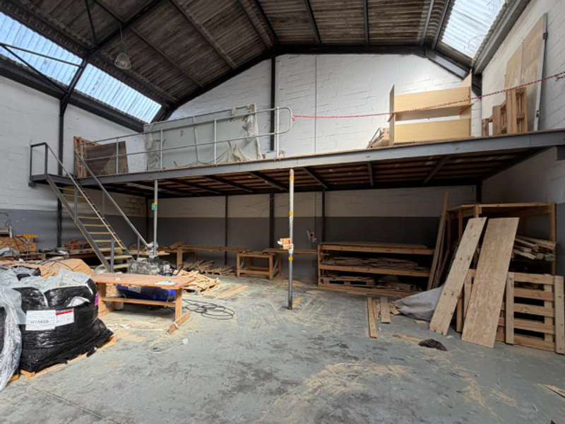 Industrial warehouse space featuring a mezzanine level, steel staircase, exposed roof trusses, and durable concrete flooring, suitable for storage or workshop use.