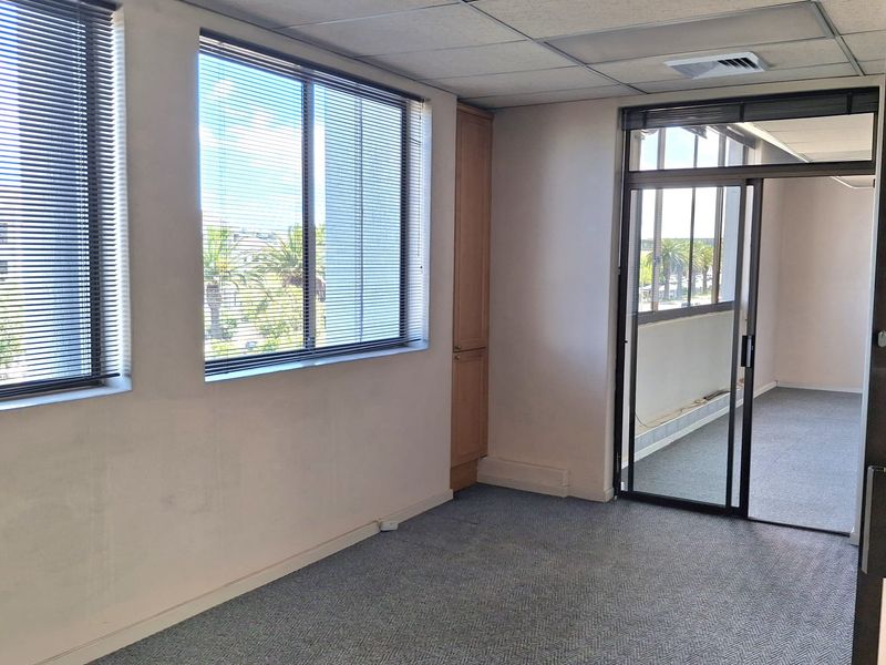 A vacant office space featuring a suspended ceiling, built-in storage cabinetry, carpeted flooring, and large windows with venetian blinds, leading to an adjacent office area.
