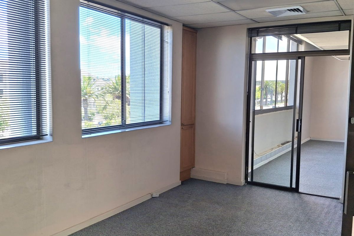 A vacant office space featuring a suspended ceiling, built-in storage cabinetry, carpeted flooring, and large windows with venetian blinds, leading to an adjacent office area.