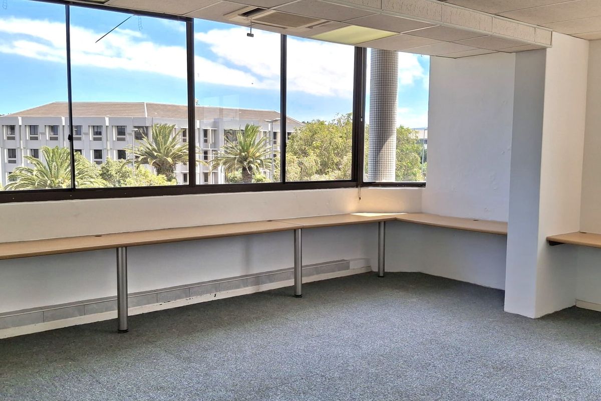An empty office space featuring large windows, built-in desk surfaces, suspended ceilings, and carpeted flooring, offering a bright and functional workspace.