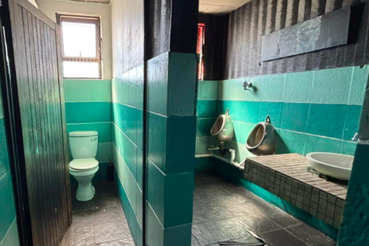 A dated commercial restroom facility featuring wall-mounted urinals, a private toilet stall, and a tiled vanity area with a vessel sink.