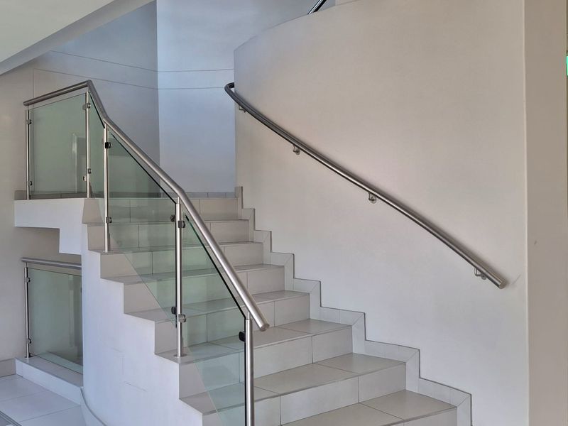 A modern, well-maintained staircase featuring tiled steps, a sleek glass balustrade, and a stainless steel handrail in a bright, minimalist office hallway.