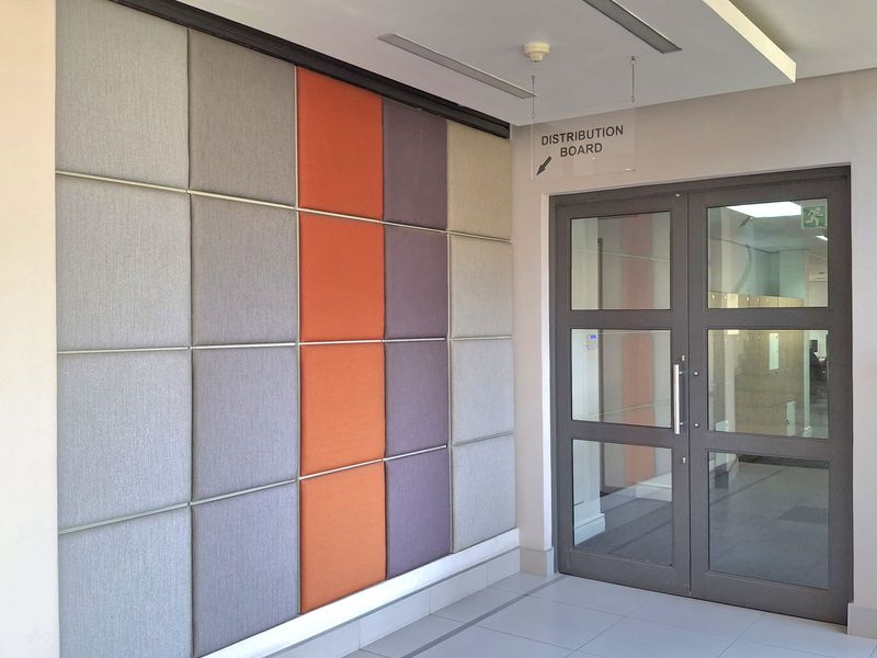 A modern office hallway featuring acoustic wall paneling, recessed ceiling lighting, and glass panel double doors leading to an adjacent workspace.