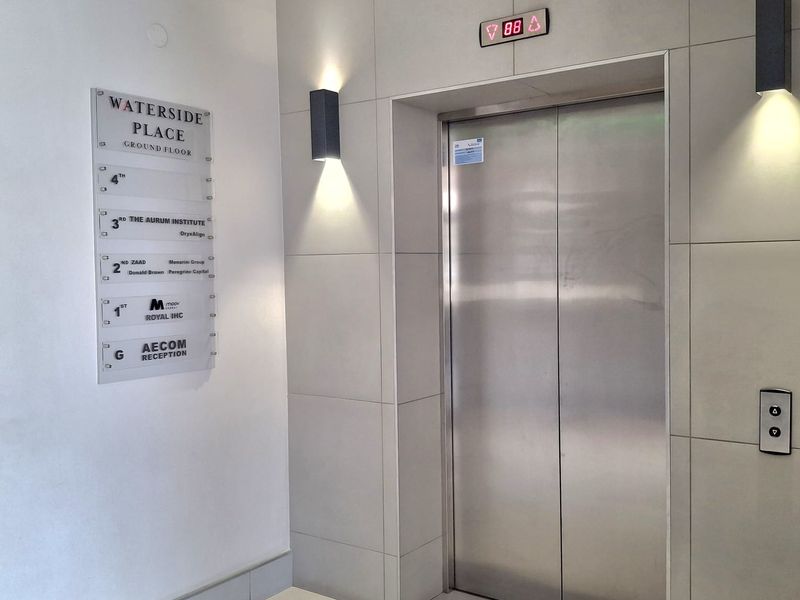 A modern office building lobby featuring a stainless steel elevator, tiled wall finishes, and a clear directory signage board for tenants.