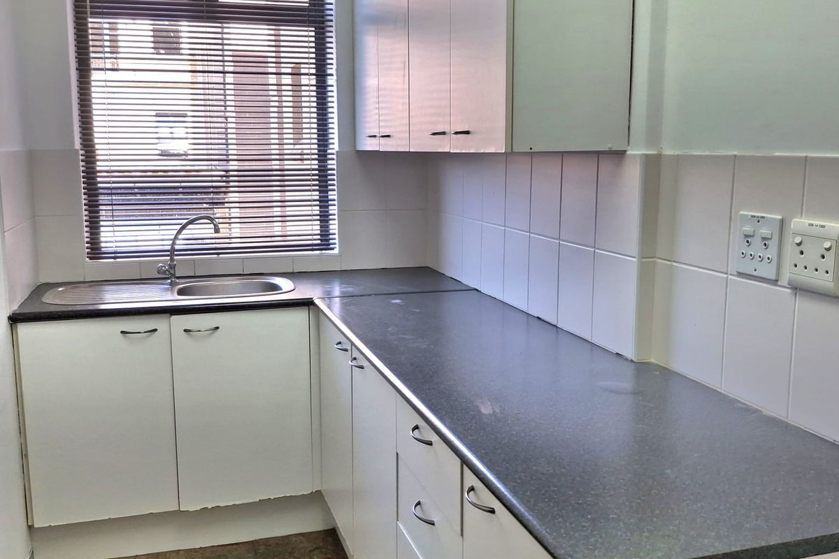 A functional office kitchenette featuring white cabinetry, dark laminate countertops, a stainless steel sink, and tiled backsplash with natural light from a window.