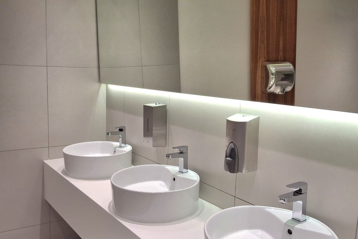 Modern office bathroom featuring a sleek vanity with three vessel sinks, wall-mounted faucets, large mirror with integrated LED lighting, and contemporary tiled finishes.