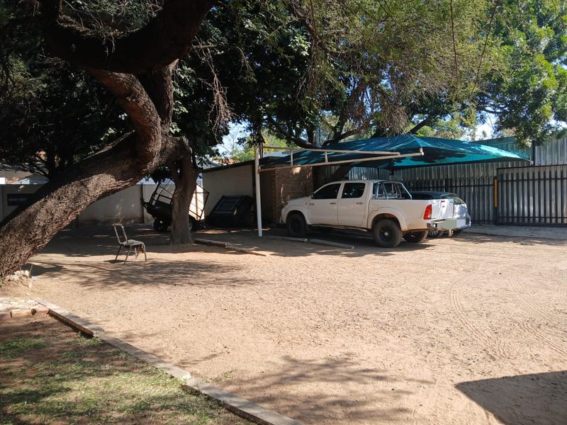 Outdoor parking area featuring a shade cloth carport structure, metal perimeter fencing, and mature trees on an unpaved surface.