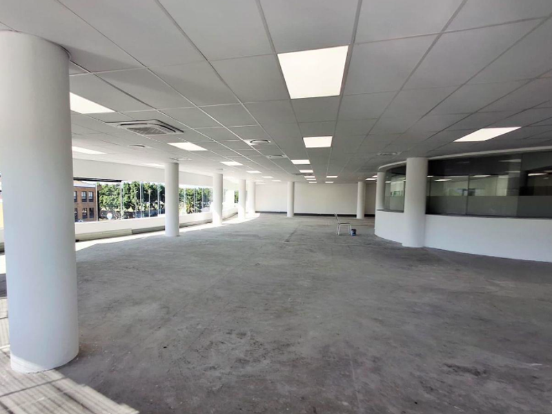 Spacious open-plan office area featuring structural columns, suspended ceilings with recessed lighting, and large windows providing ample natural light.
